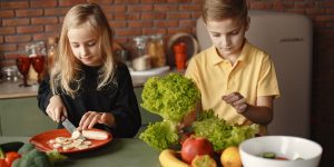 Focused little girl and boy sitting at desk with assorted vegetables and fruits while cooking in loft kitchen with brick wall