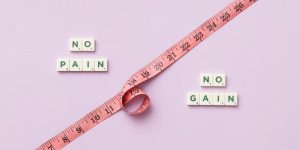 Pink tape measure with 'No Pain No Gain' spelled in Scrabble tiles on pink background.